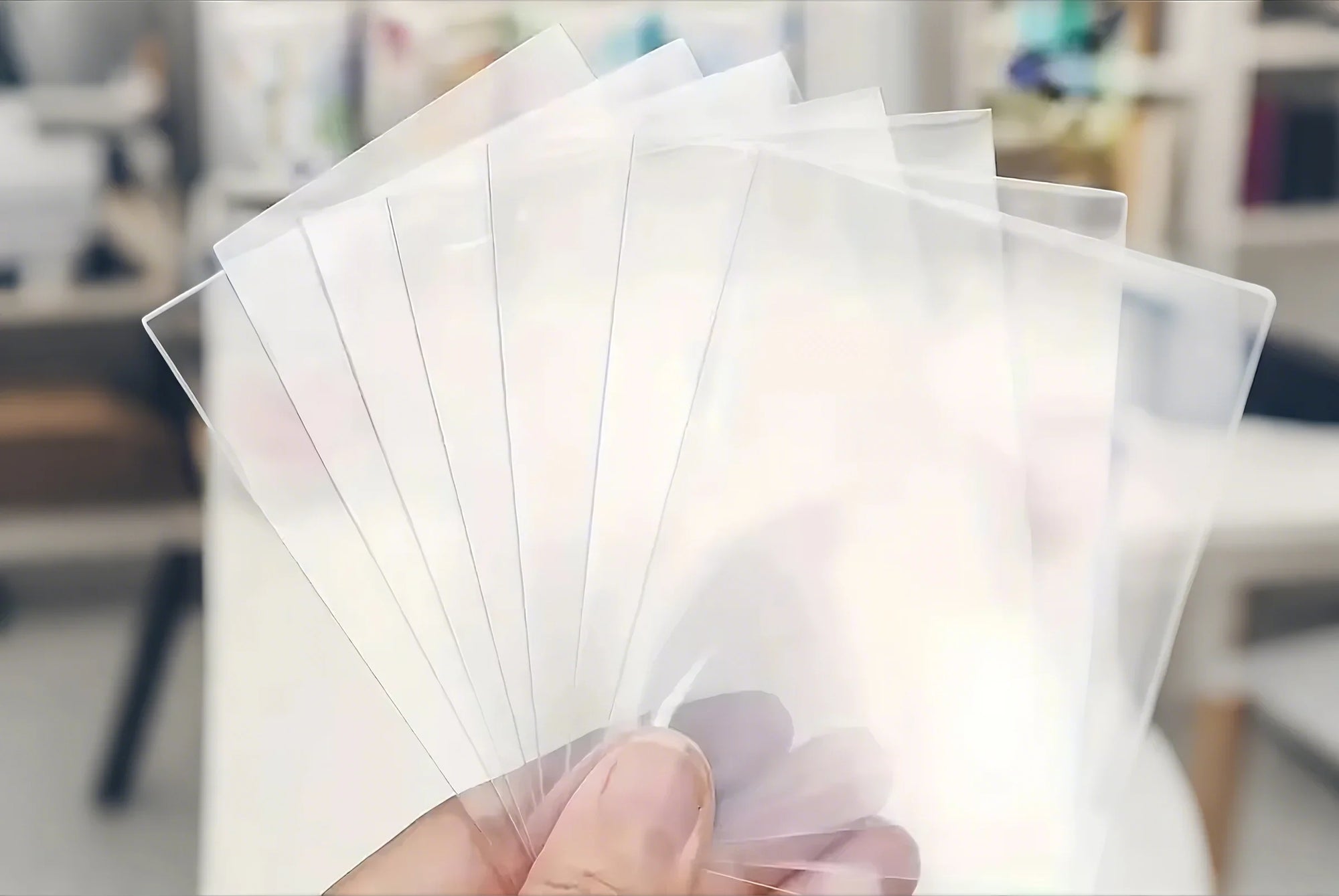 Hand holding a stack of clear plastic Penny Sleeves with a blurred indoor background